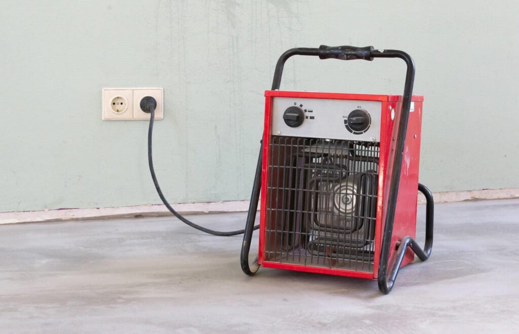 Portable red electric heater plugged into a wall outlet on a concrete floor.